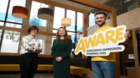 Three people standing inside a Danske Bank branch smiling at the camera. One is holding a large sign with the yellow AWARE logo on it.