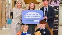 Young Enterprise NI - May 2022 Three adults and two school pupils standing in a shopping mall. They are holding a small sign that says 'Young Enterprise NI Business Beginnings, Powered by Danske Bank'.